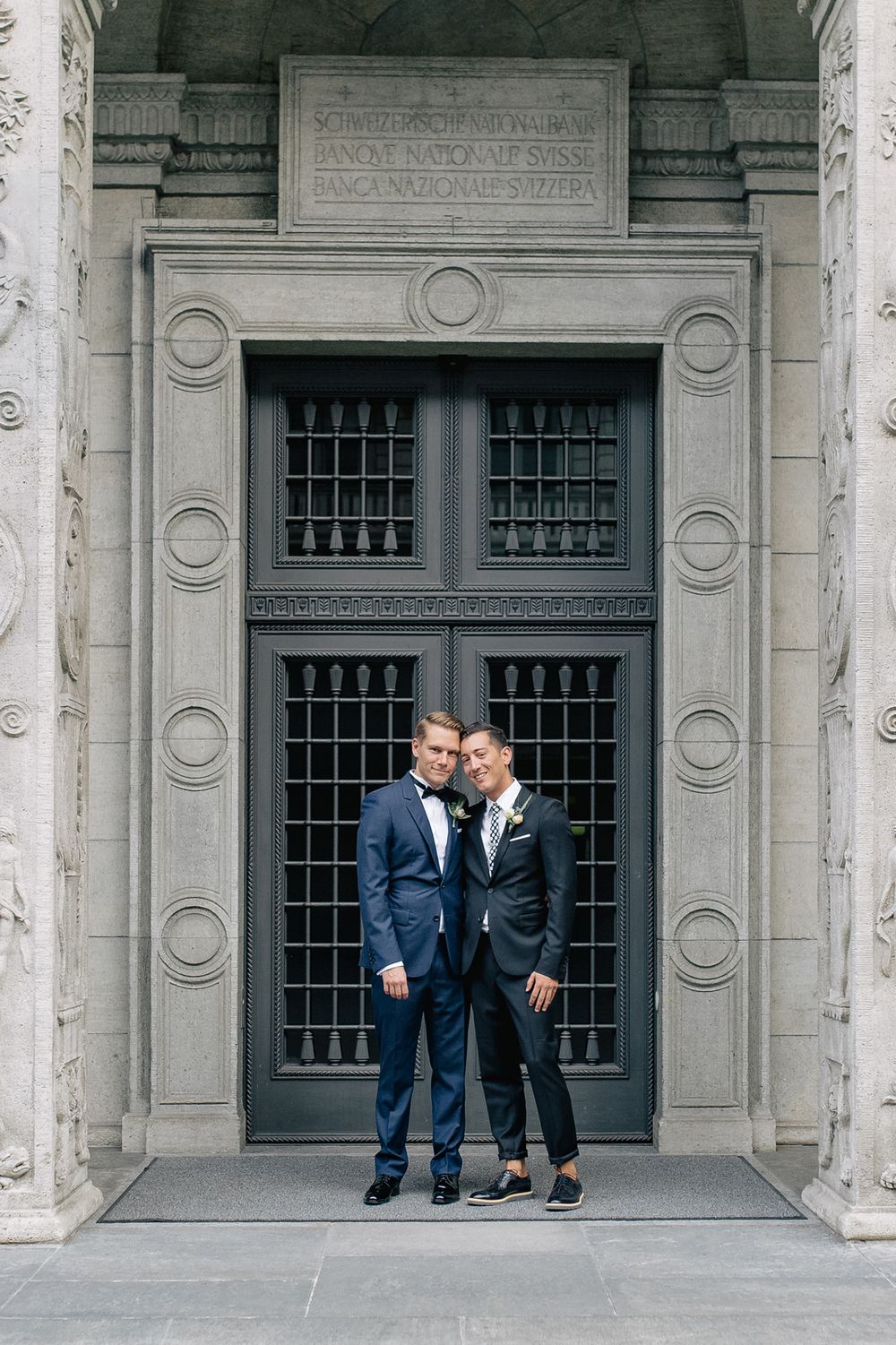A couple in matching blue suits standing before a large, symmetrical stone doorway at a historic Zurich venue.