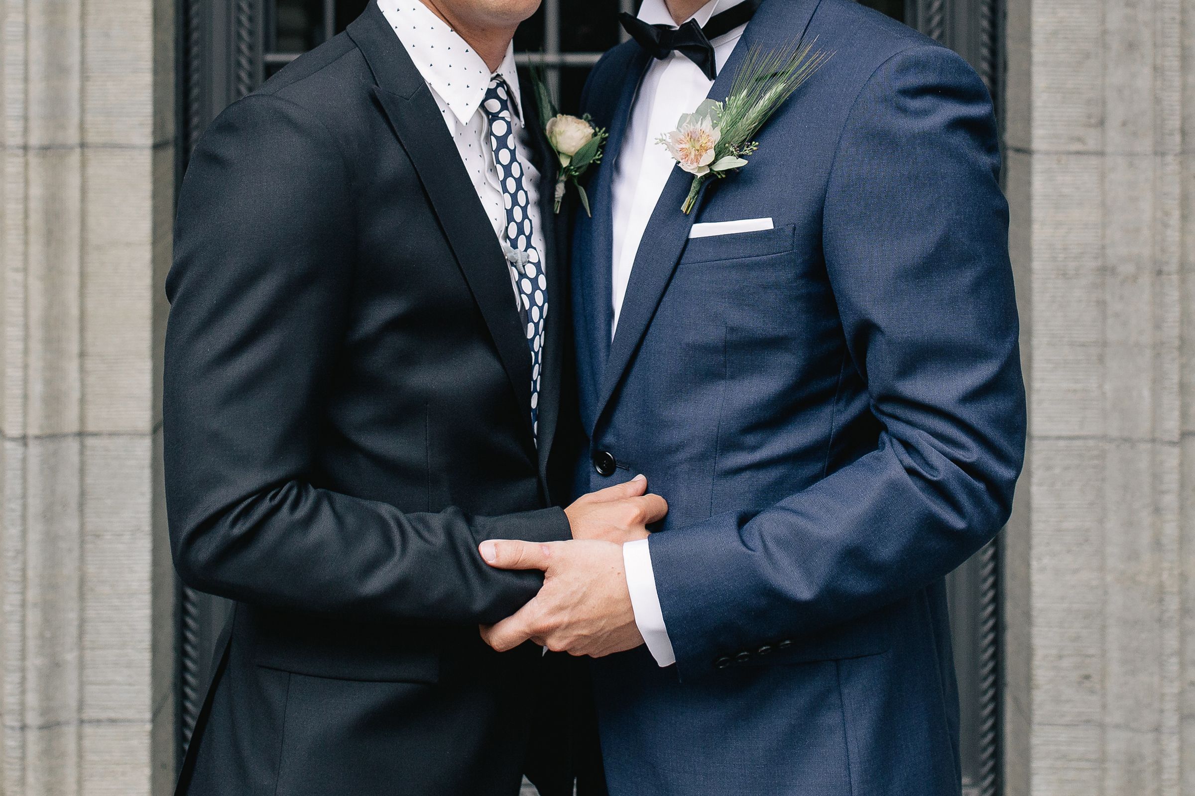 Close-up portrait of two grooms standing together, showcasing their dark blue wedding suits and floral boutonnieres.