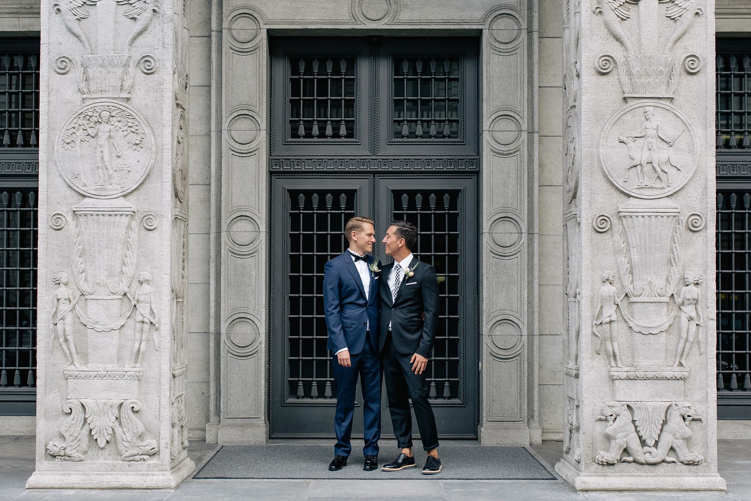 Wide shot of two grooms in blue suits posing in front of a grand stone entrance with ornate carvings.