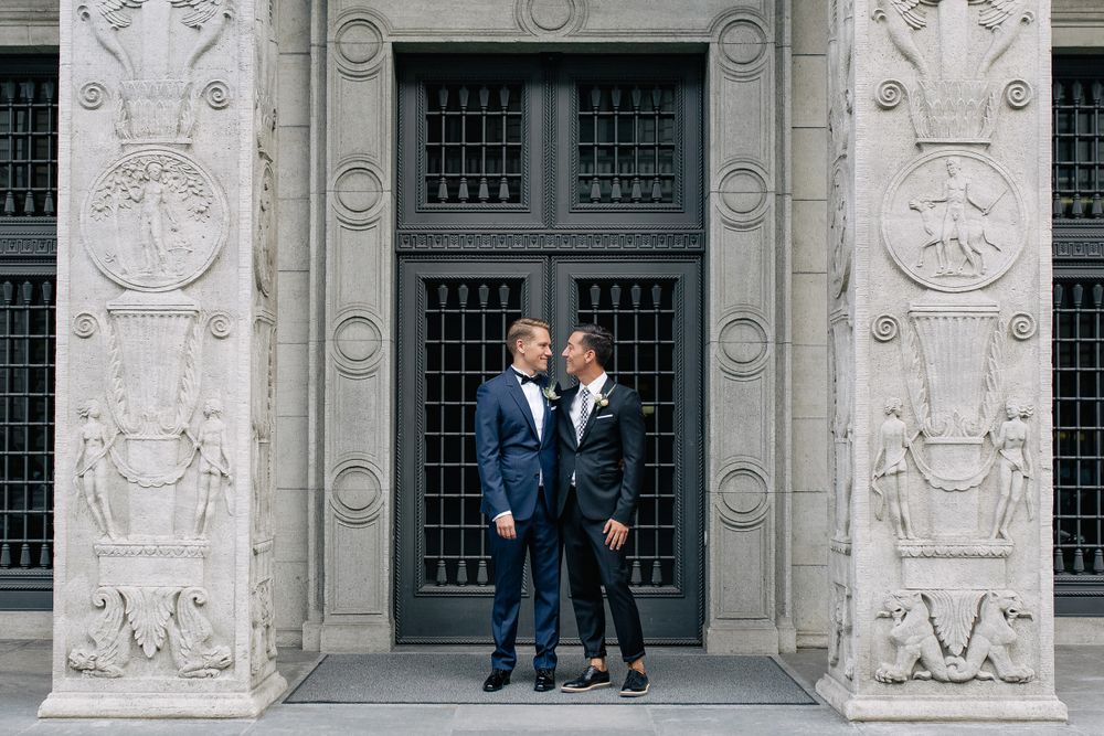 Wide shot of two grooms in blue suits posing in front of a grand stone entrance with ornate carvings.