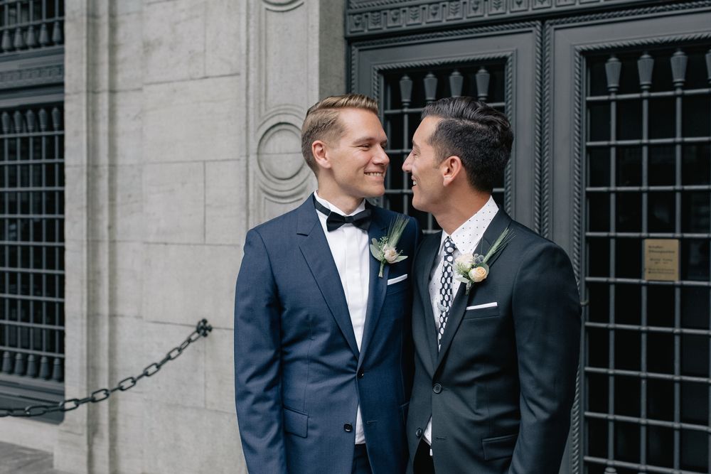 Horizontal portrait of two grooms smiling together in front of a historic building's exterior.