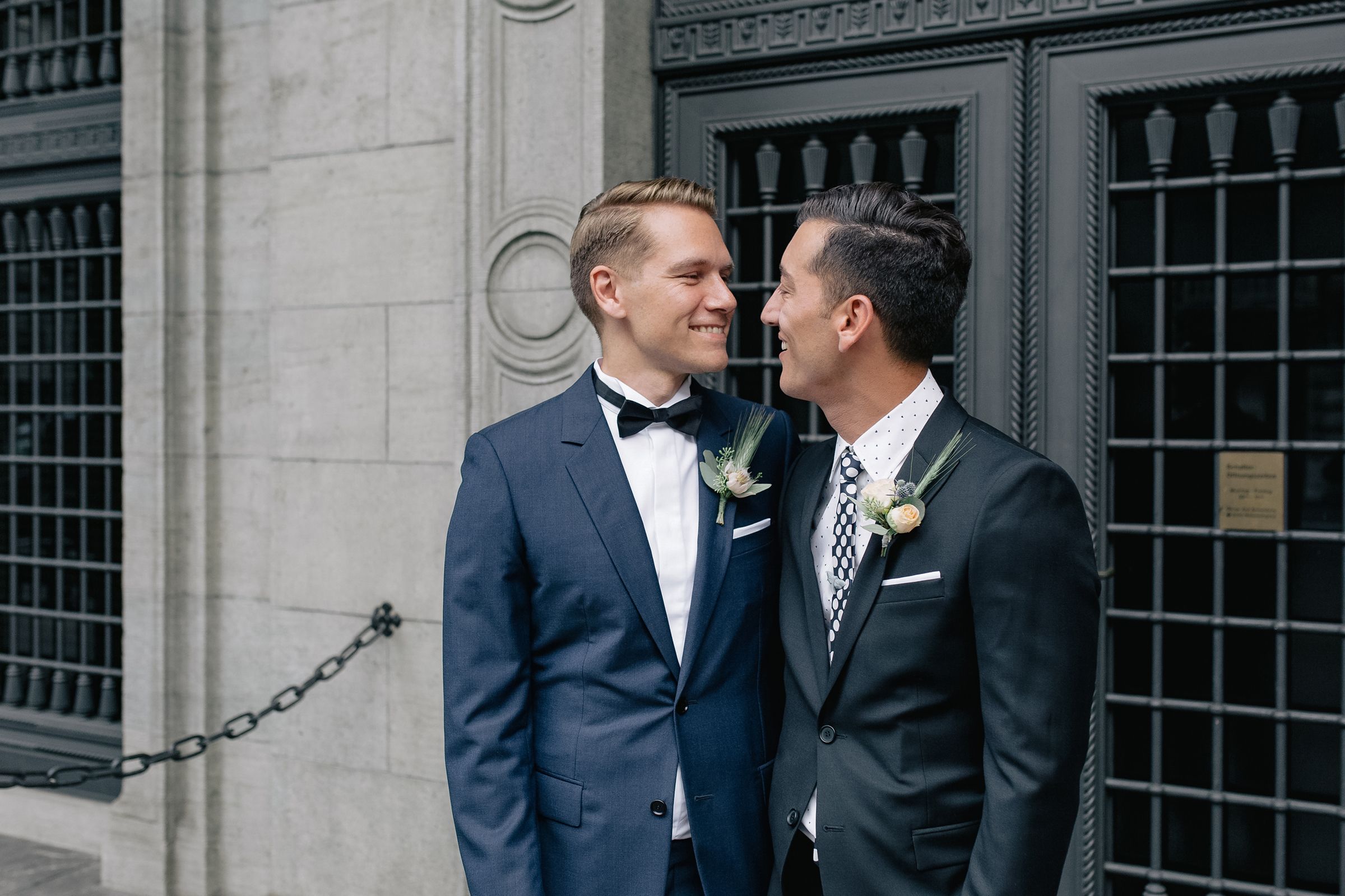Horizontal portrait of two grooms smiling together in front of a historic building's exterior.