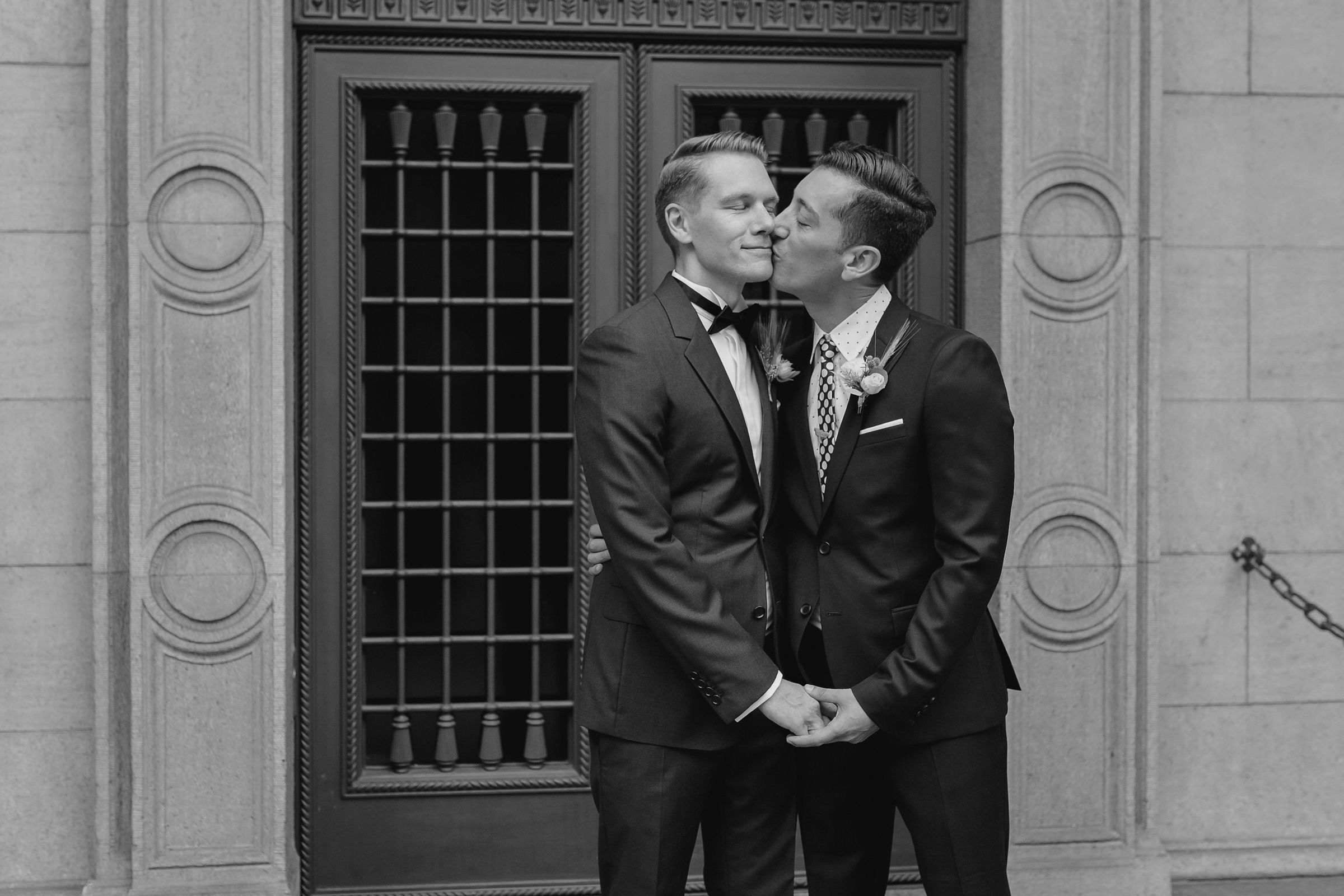 Black and white close-up of a groom kissing his partner's cheek in front of a lattice door.