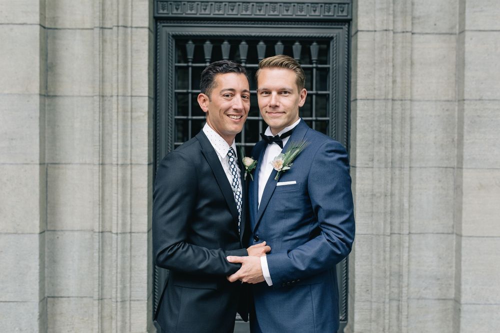 Horizontal portrait of two grooms smiling together in front of a historic building's exterior.