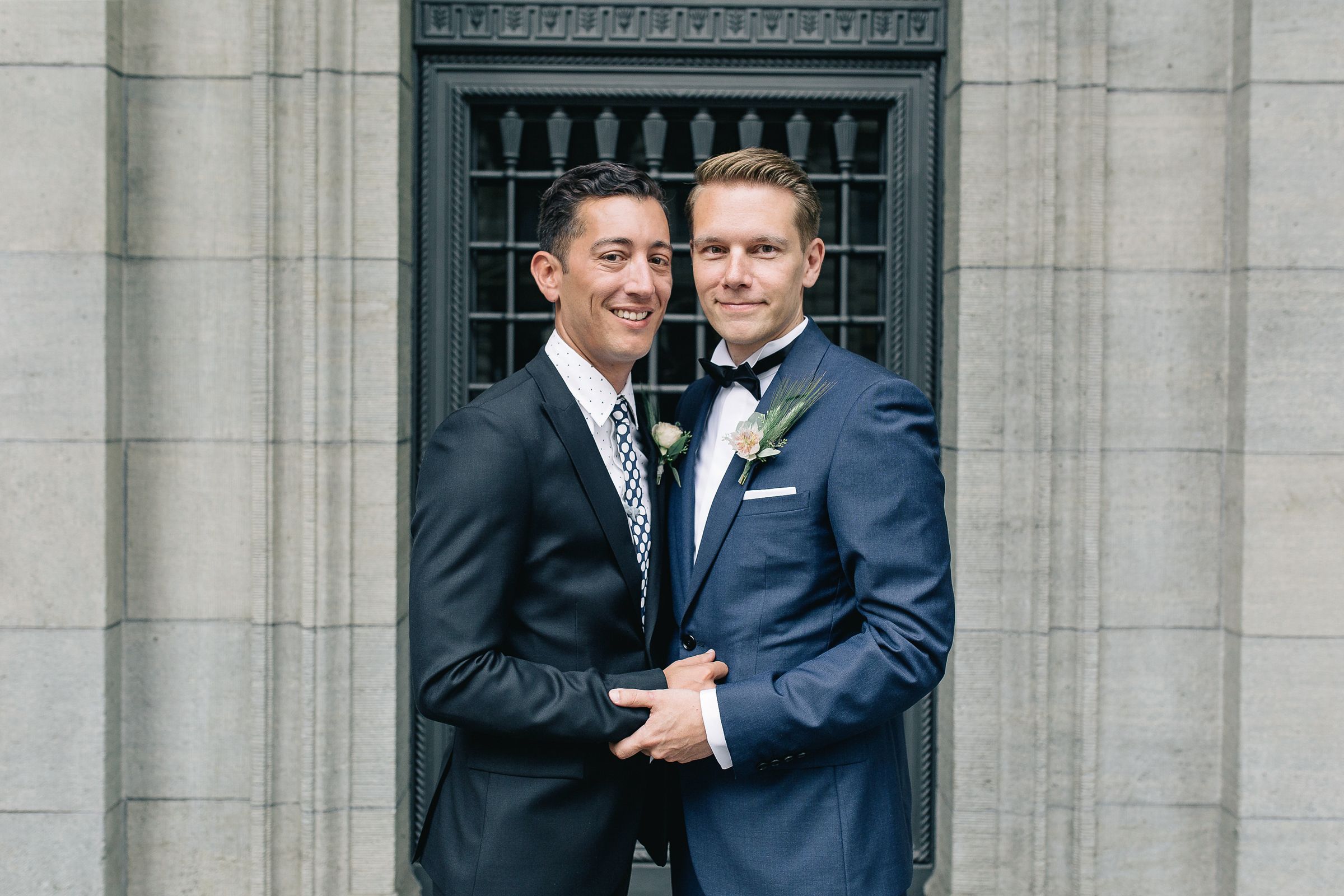 Horizontal portrait of two grooms smiling together in front of a historic building's exterior.