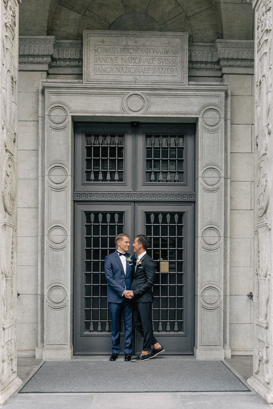 Wide architectural shot of a couple posing before a large, ornate stone-framed entrance.
