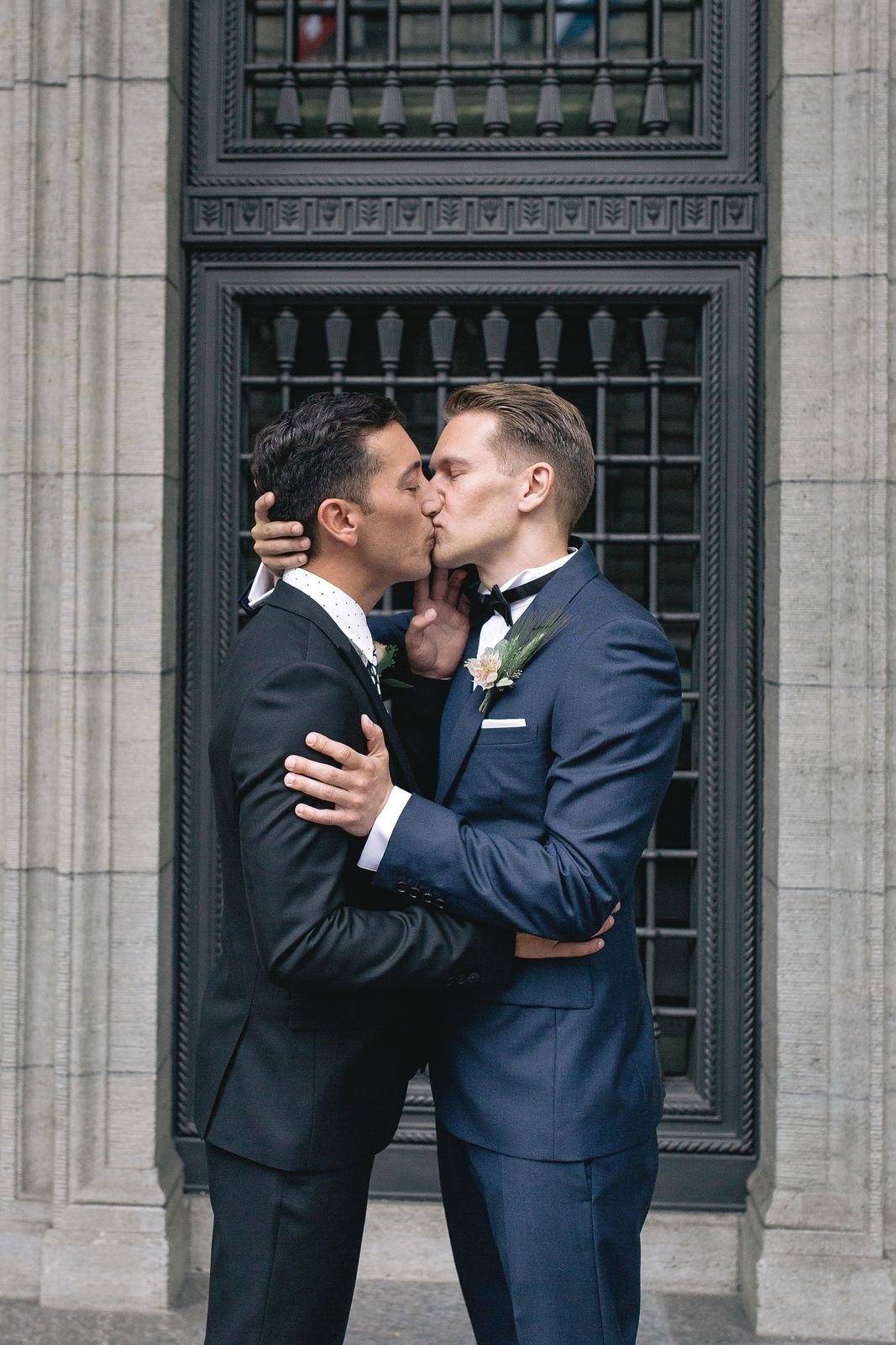 Two grooms in blue suits sharing a romantic kiss in front of a grand, dark-framed door.