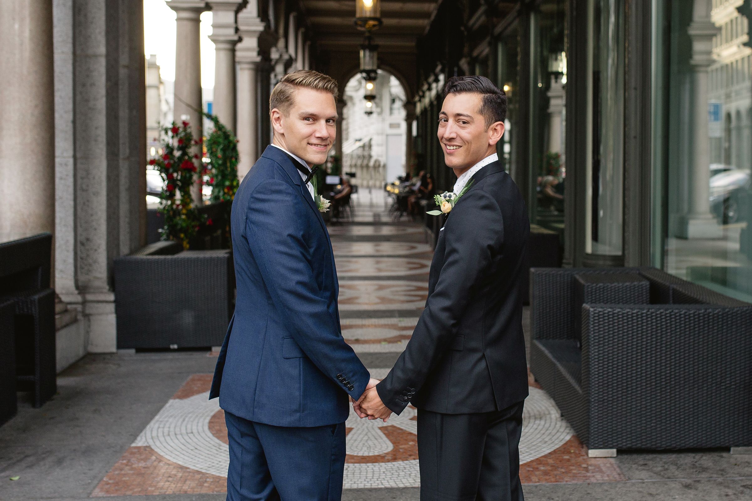 Two grooms in blue suits holding hands and standing together in an elegant, sunlit hallway.