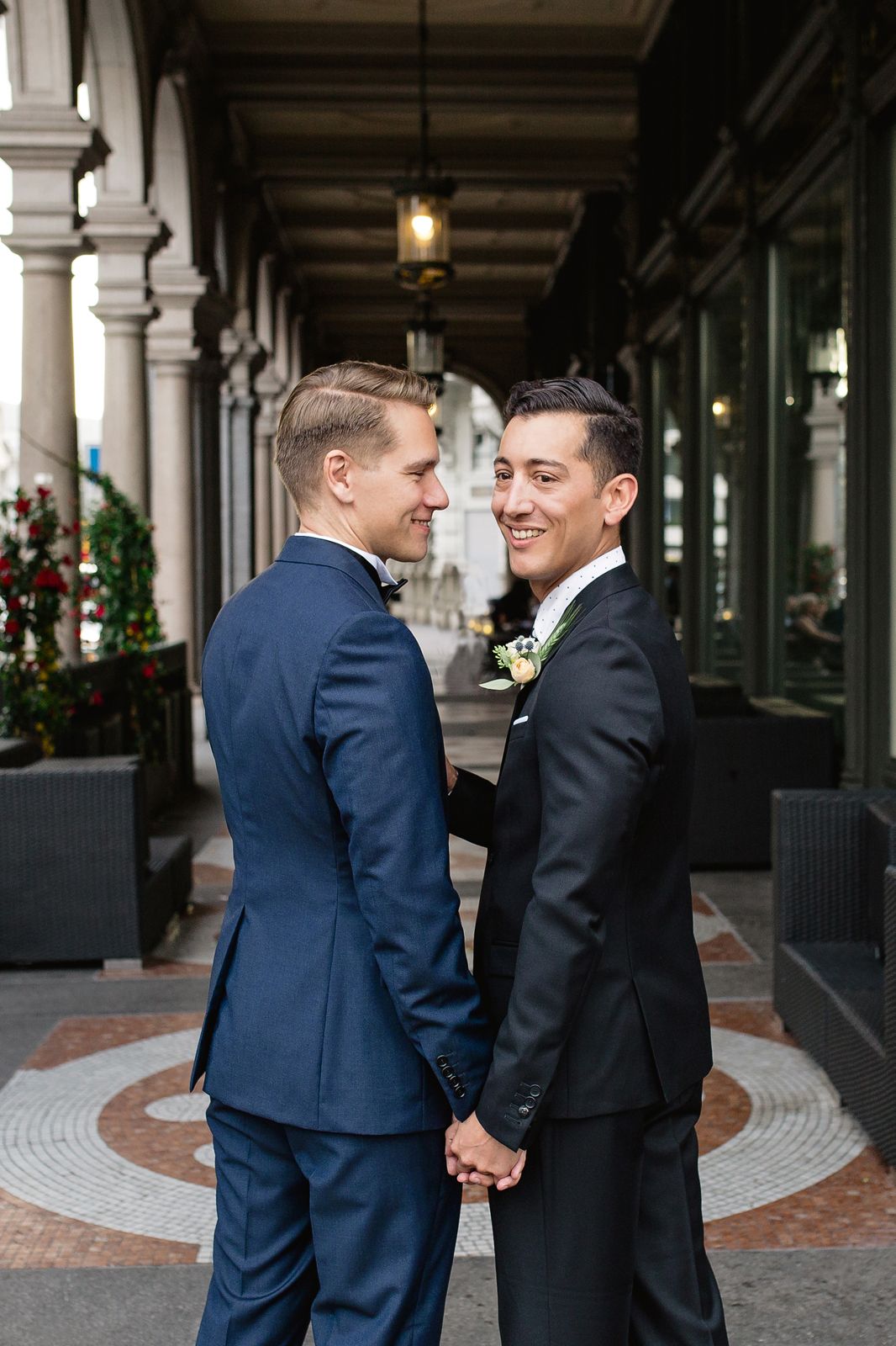 Full-length vertical shot of two grooms posing together in a modern, light-filled venue corridor.