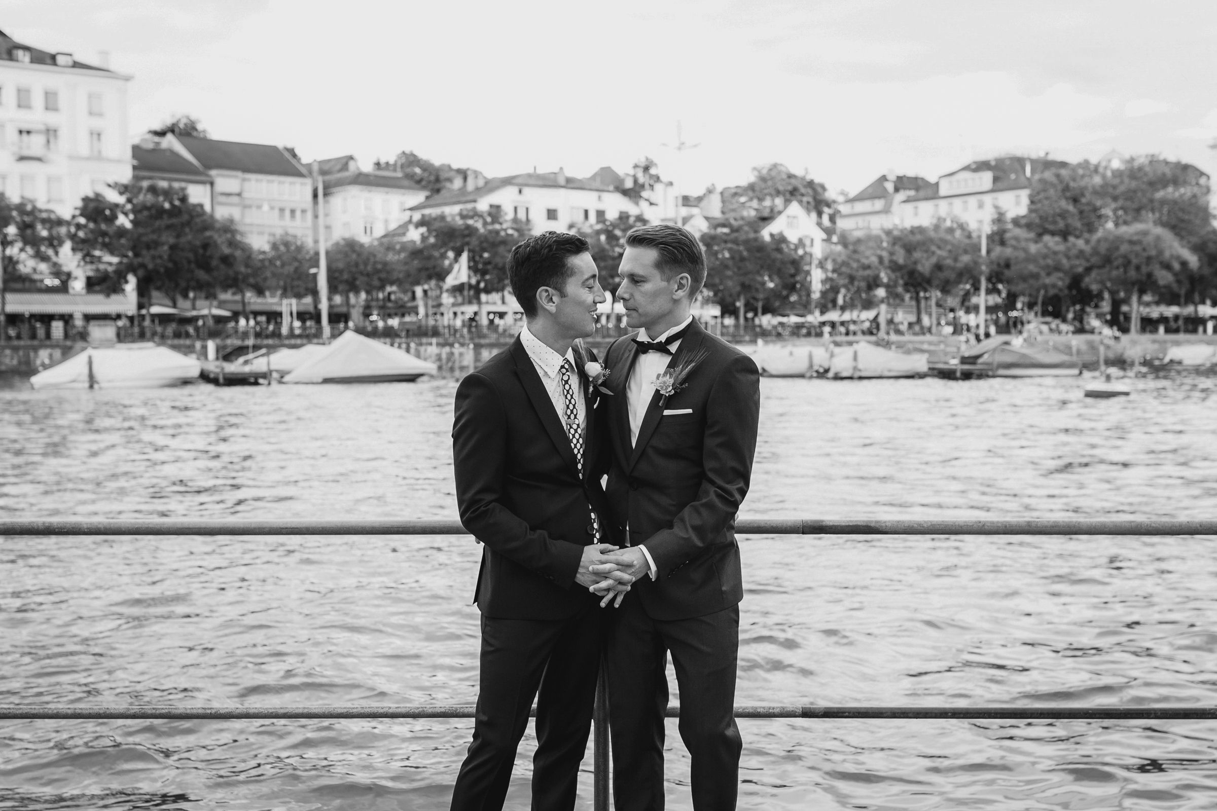 Wide black and white portrait of a guy couple holding hands on a waterfront in Zurich with boats behind them.