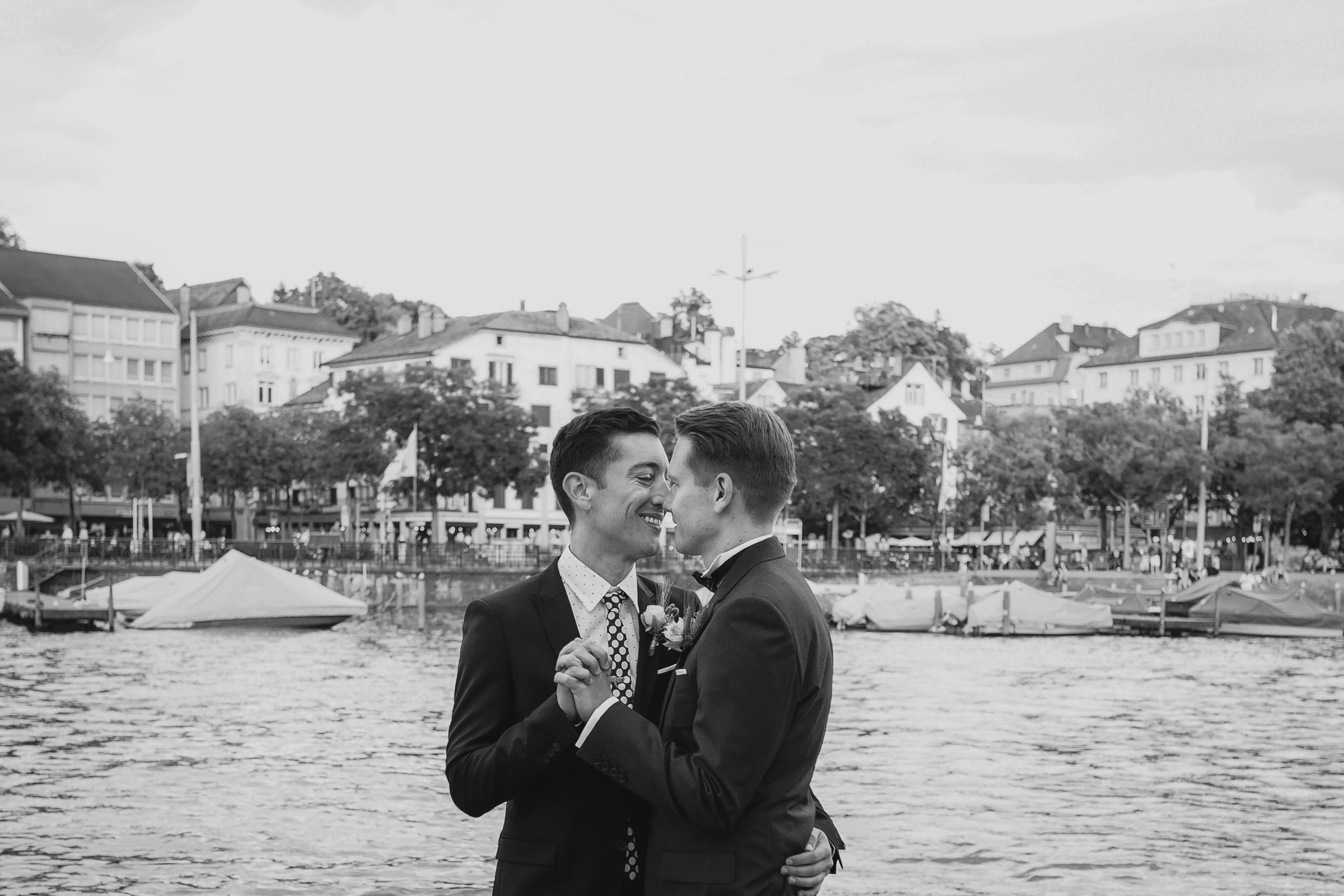 Black and white candid shot of a guy couple dancing outdoors with the Zurich skyline and river in the background.
