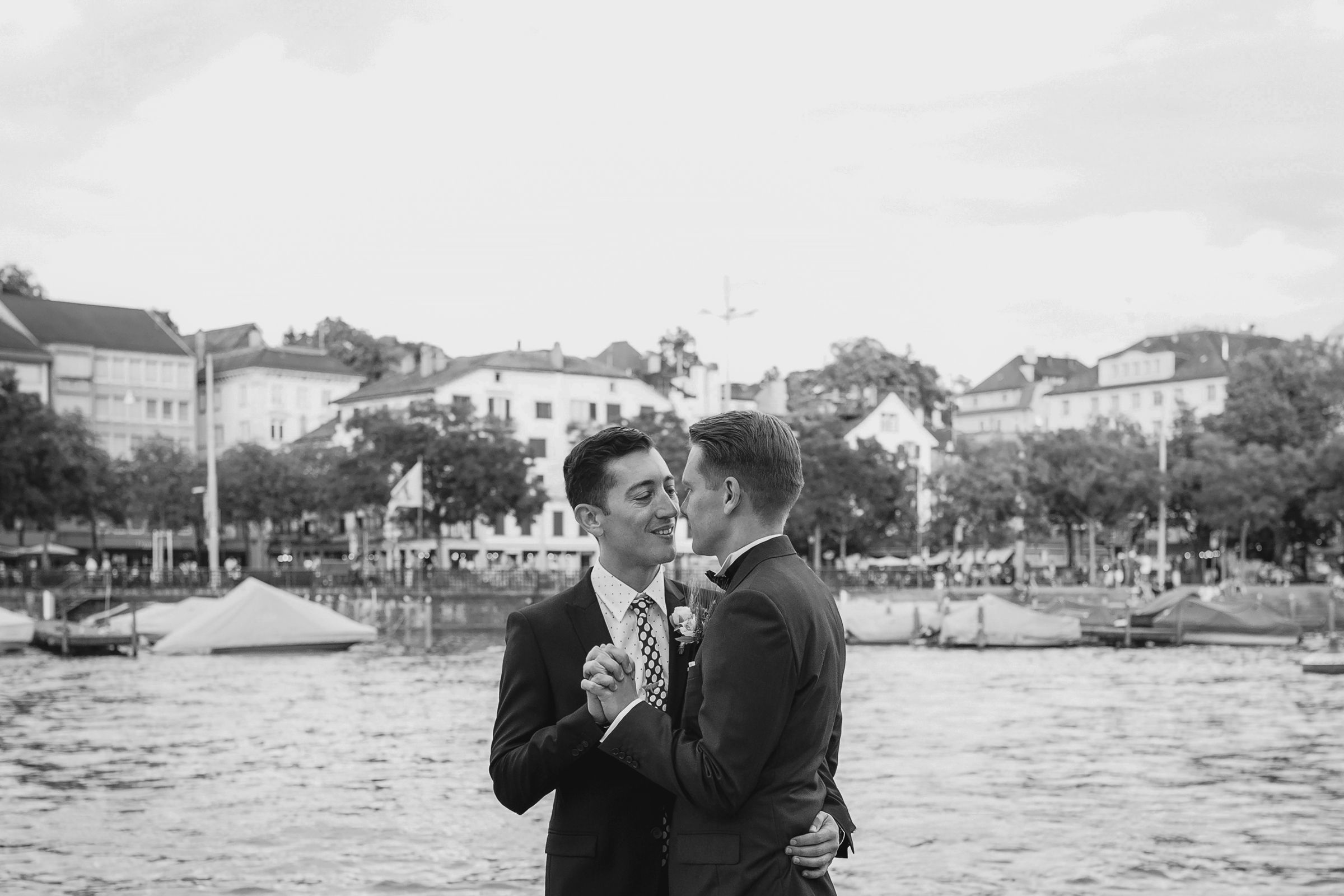 Wide black and white shot of a couple standing on a waterfront with boats and Zurich's Old Town behind them.