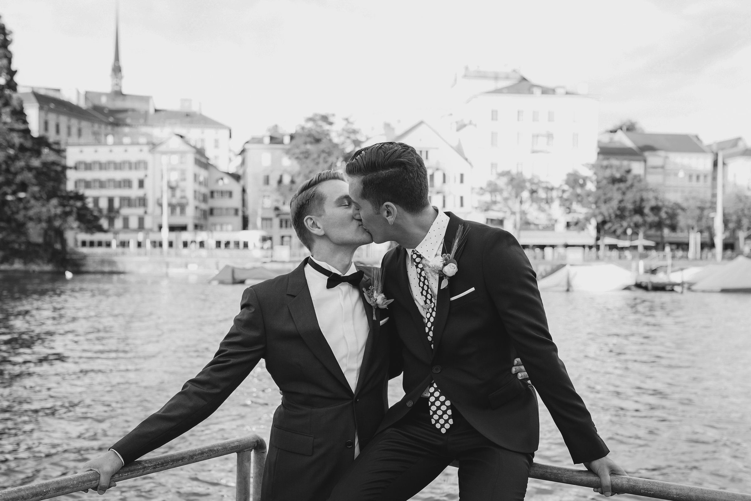 Black and white landscape photo of a groom kissing his partner on a bridge overlooking the Zurich cityscape.