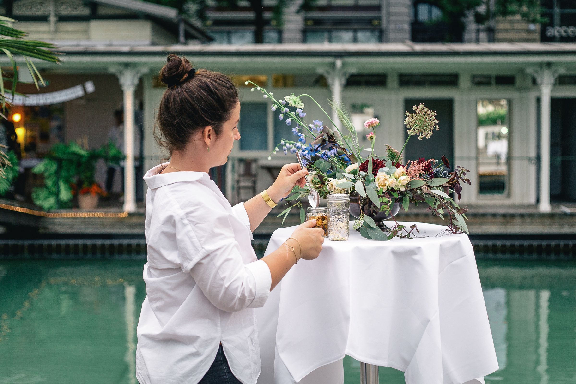 A woman setting up floral arrangements on a poolside table at an outdoor wedding reception.