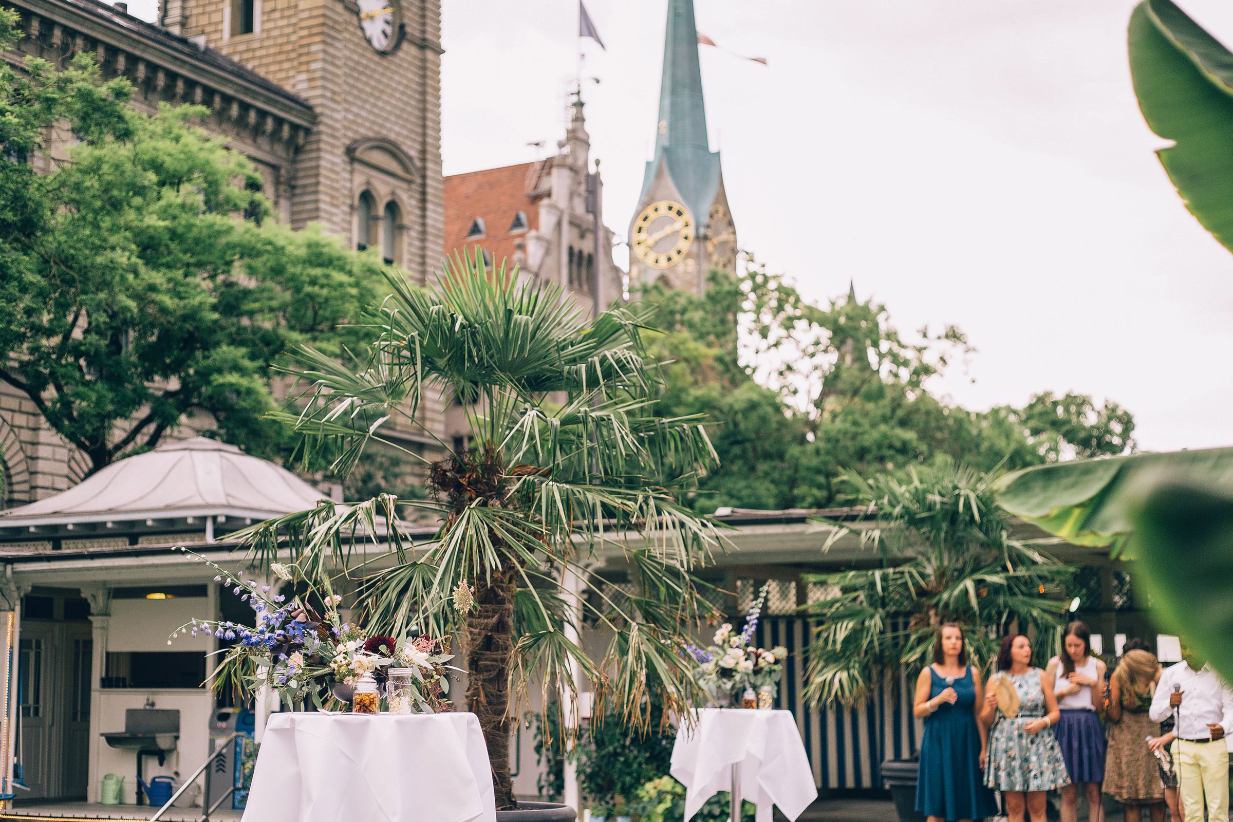 Wide shot of an outdoor wedding reception area with guests mingling under palm trees and historic city views.