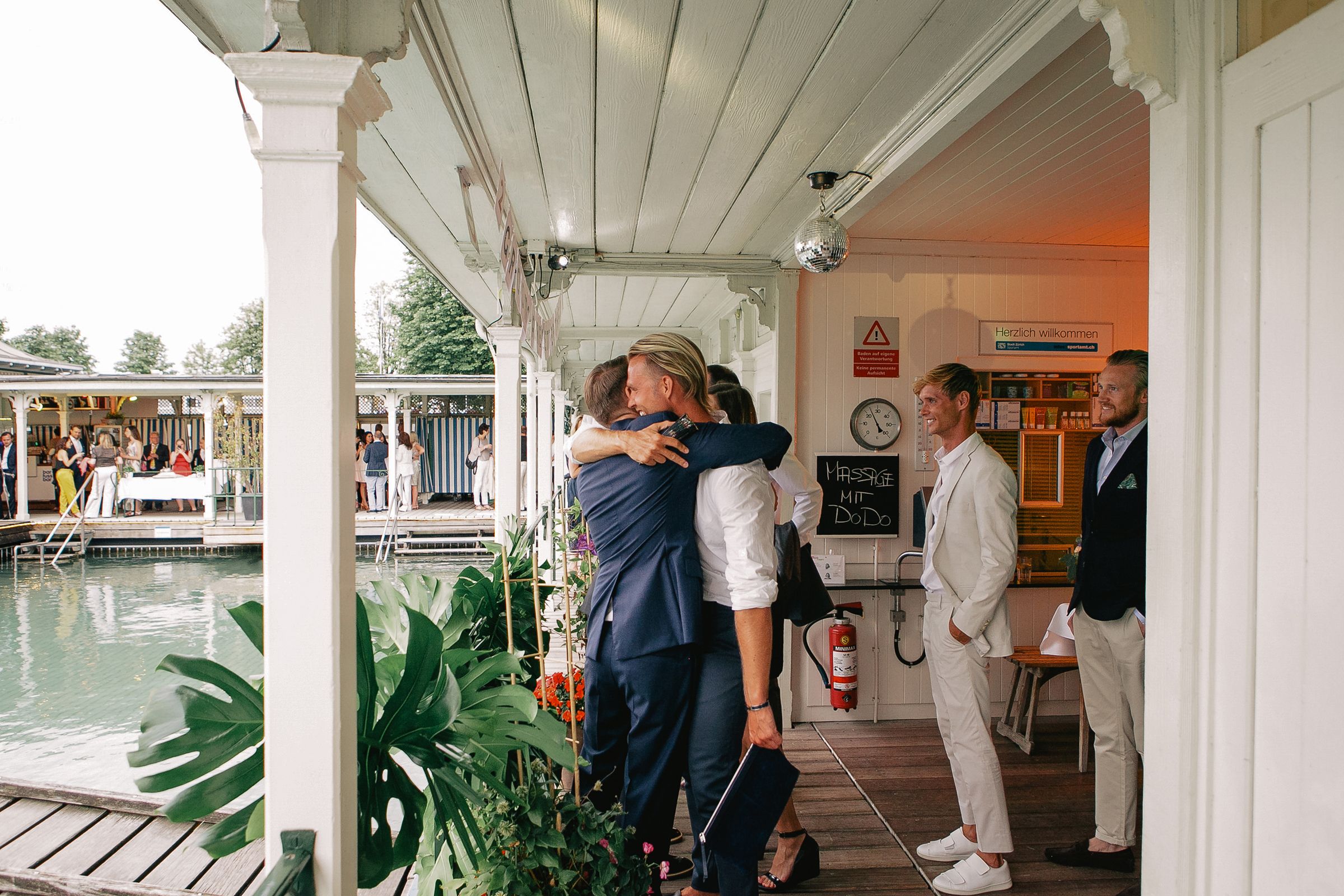 Candid shot of two men sharing a friendly hug on a white-pillared porch during a wedding reception.