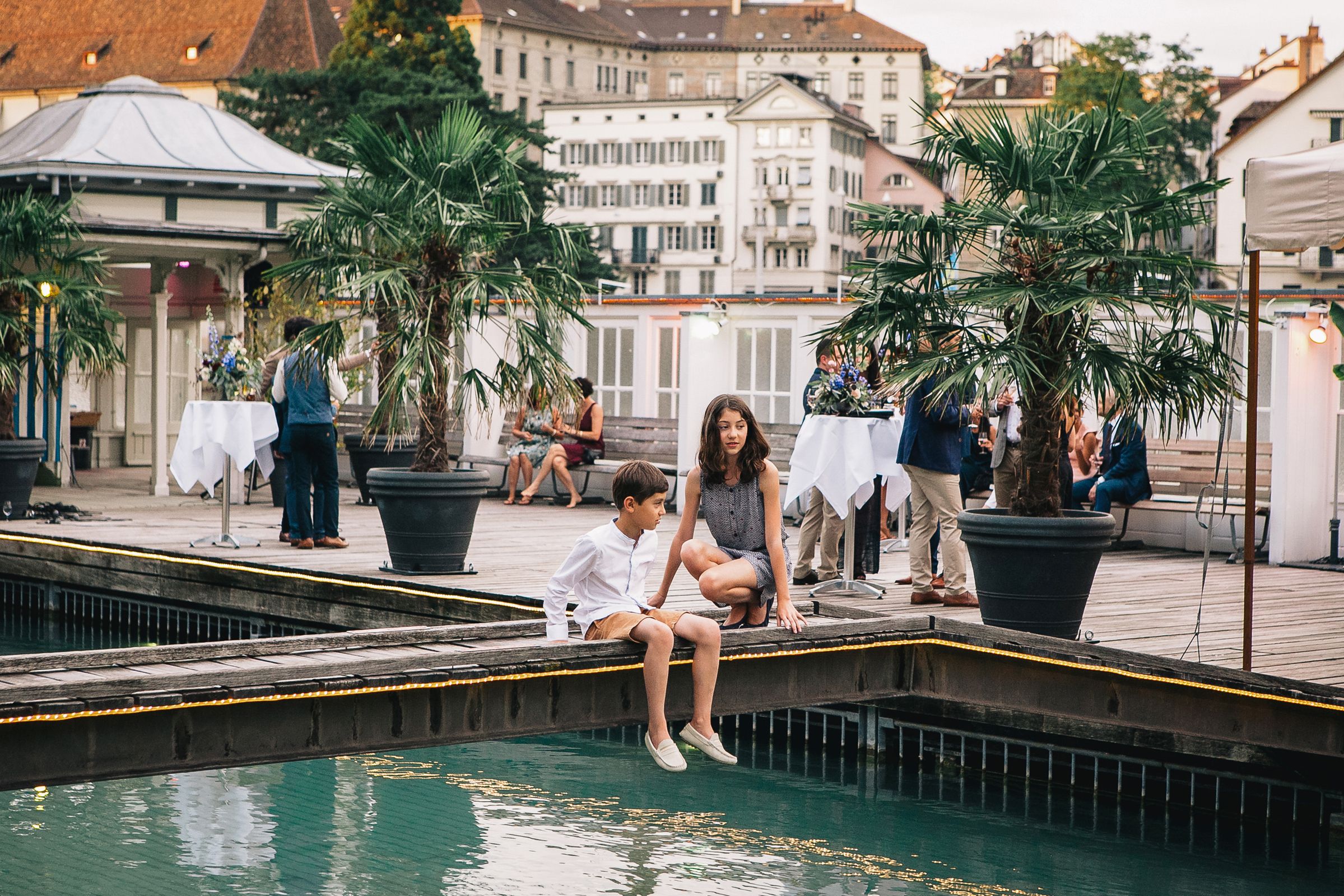 A boy and a girl relaxing on a dock, overlooking a building, capturing a peaceful moment by the water.