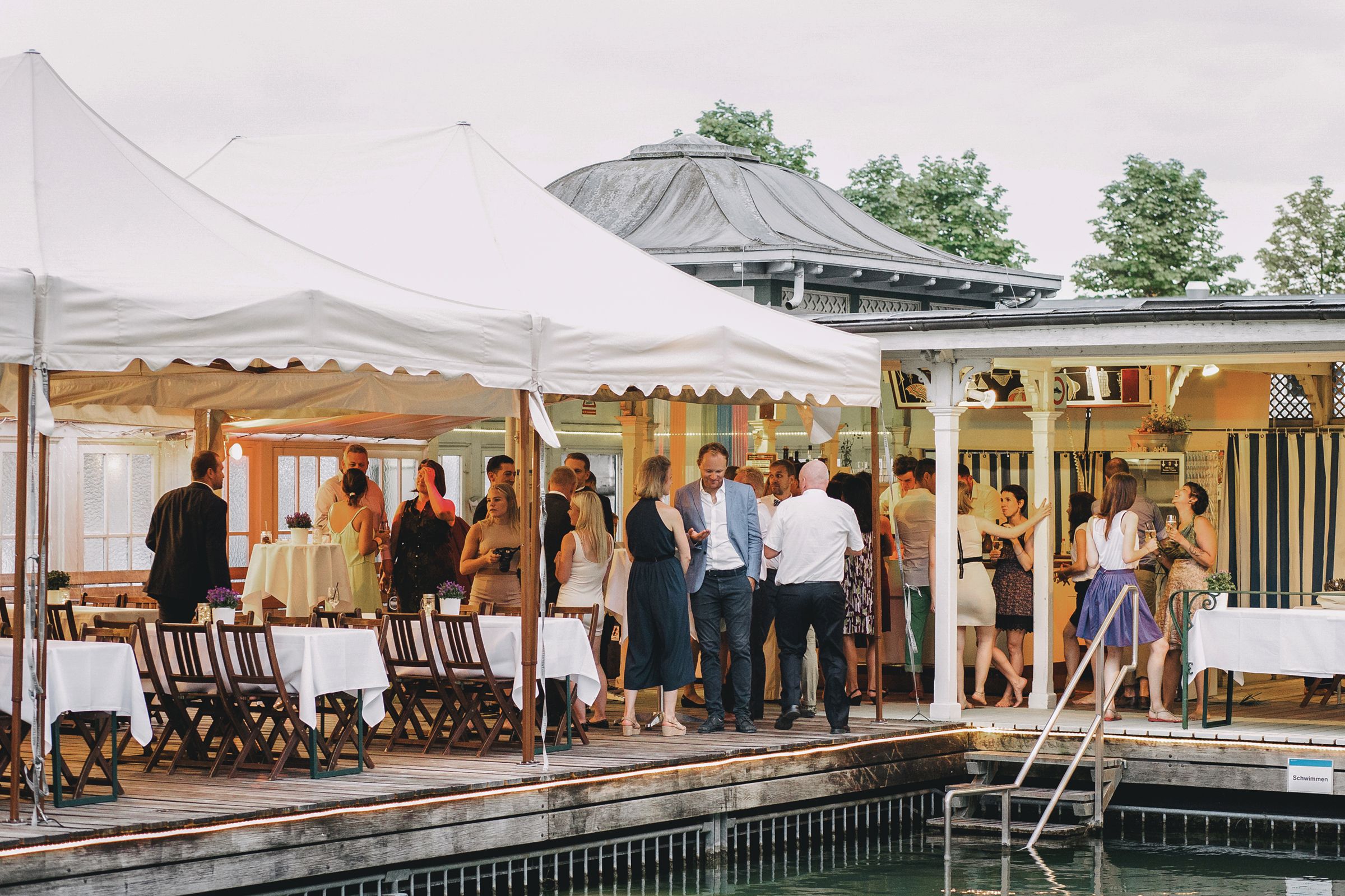 Wide shot of guests mingling outside a white marquee tent by a pool at an outdoor wedding venue.