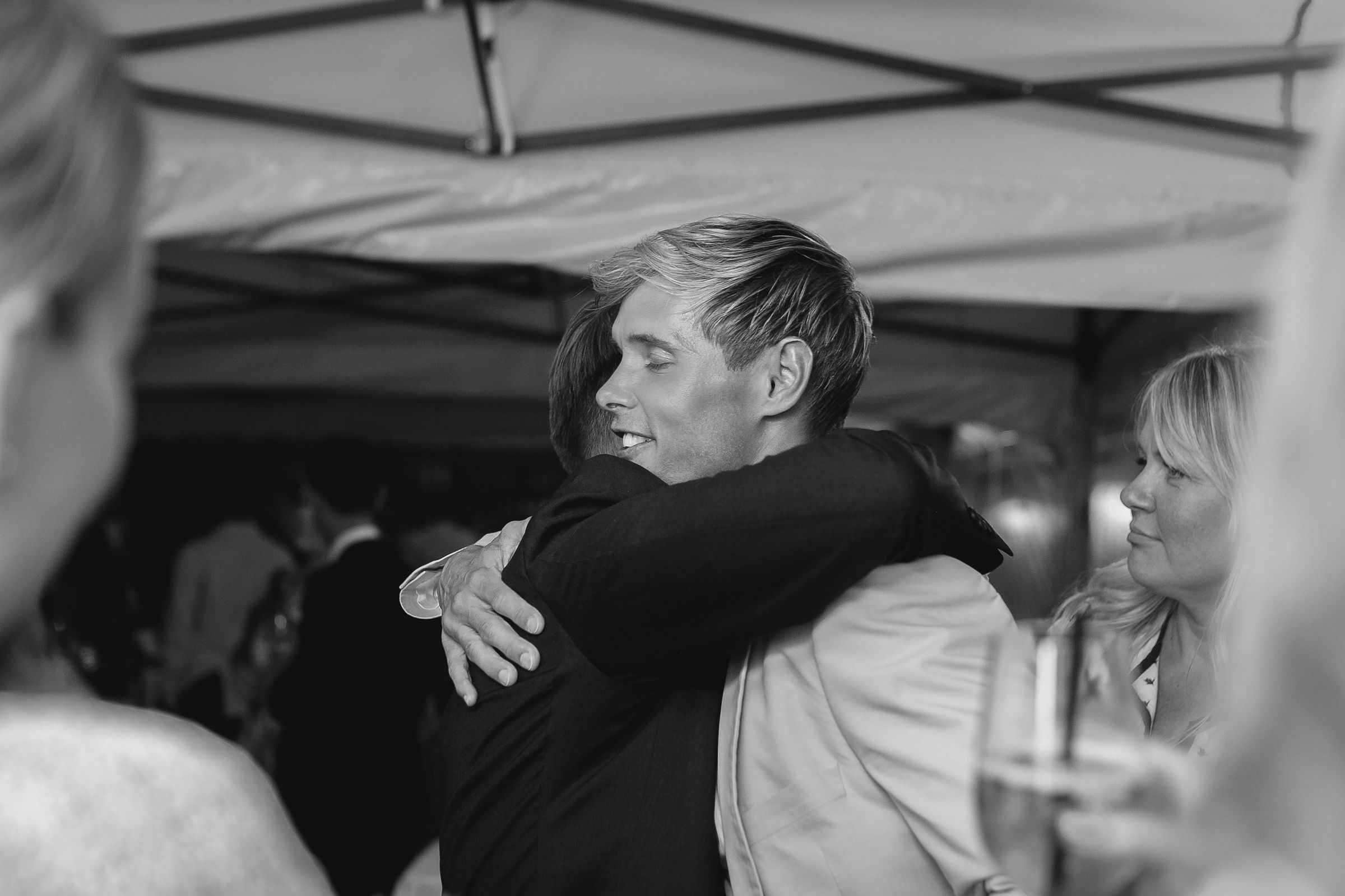 Black and white candid photo of a groom hugging a guest at an outdoor wedding reception.