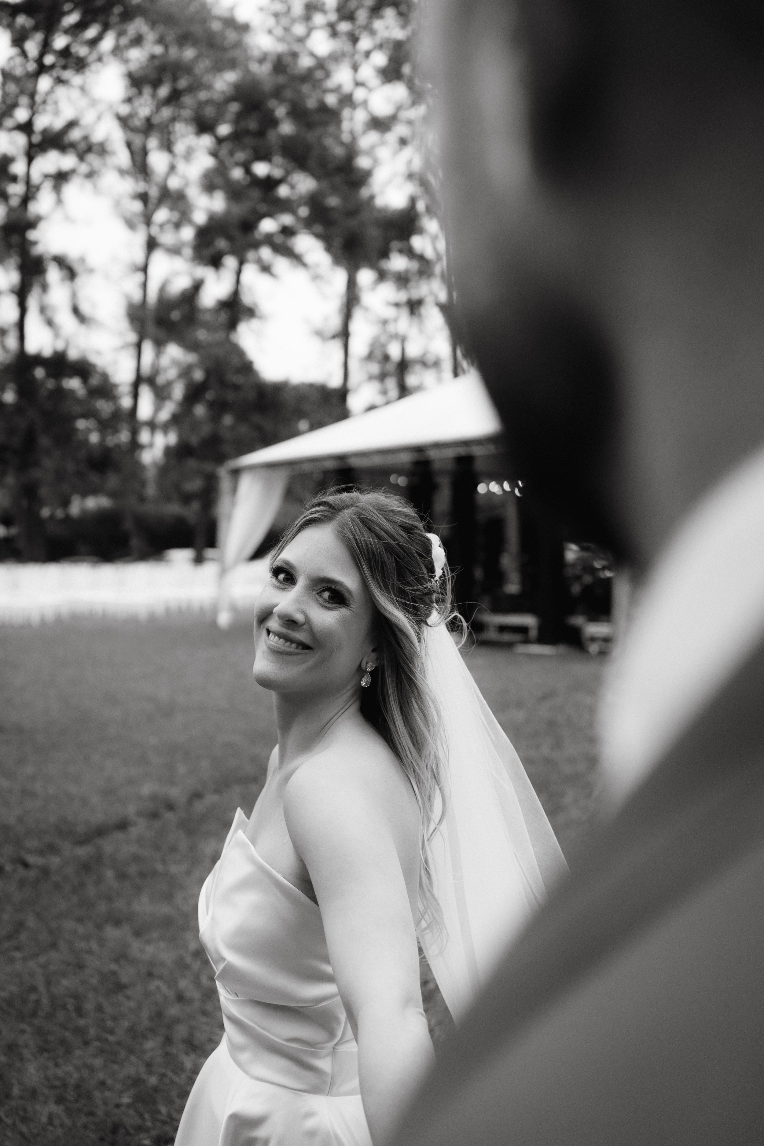 A bride in a white dress poses with a joyful expression during an outdoor wedding ceremony.