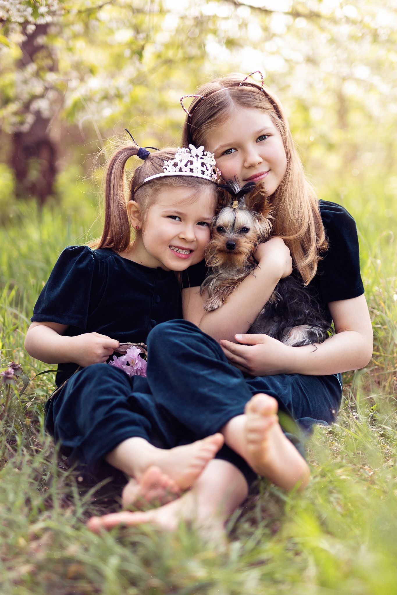 Two children sitting in grass cuddling with a small dog on a sunny afternoon.