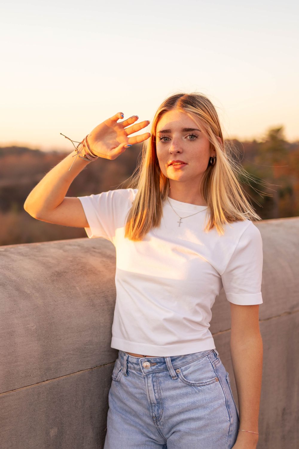 Person in white t-shirt shields eyes from sunlight while posing against concrete wall at sunset.
