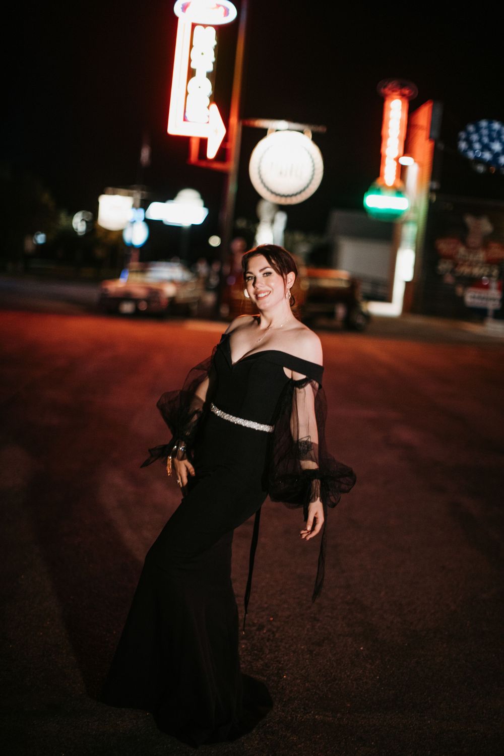 Bride in stunning black off-shoulder gown with tulle sleeves posing near neon signs at night.