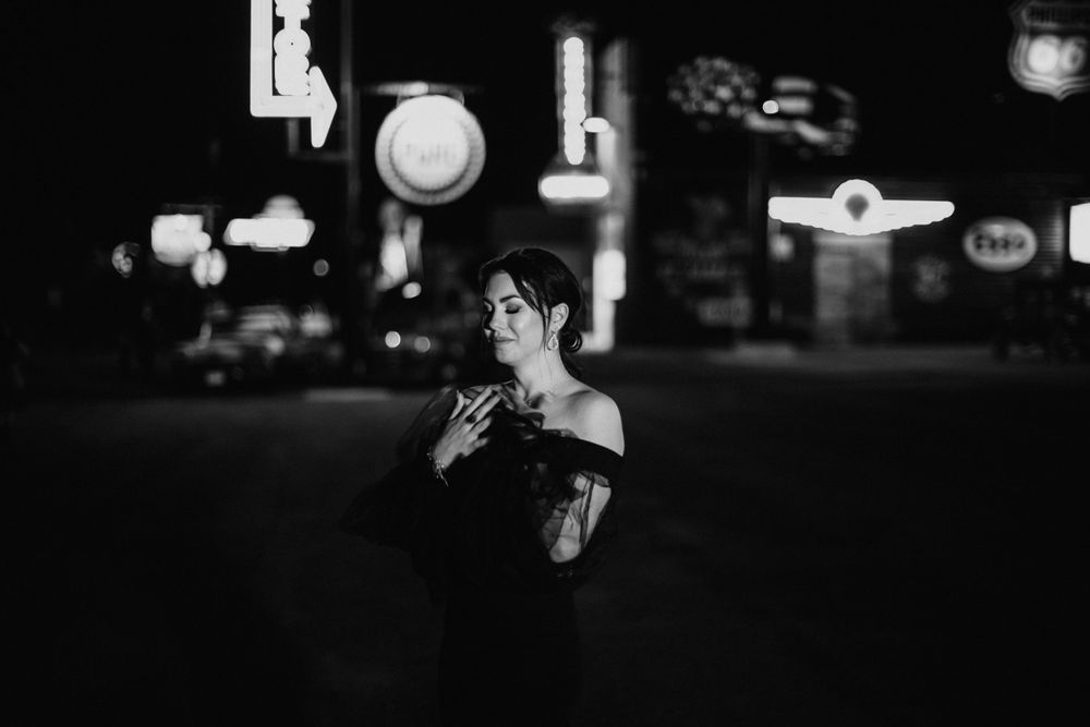 Elegant woman in black off-shoulder gown posing on a city street at night with neon signs.