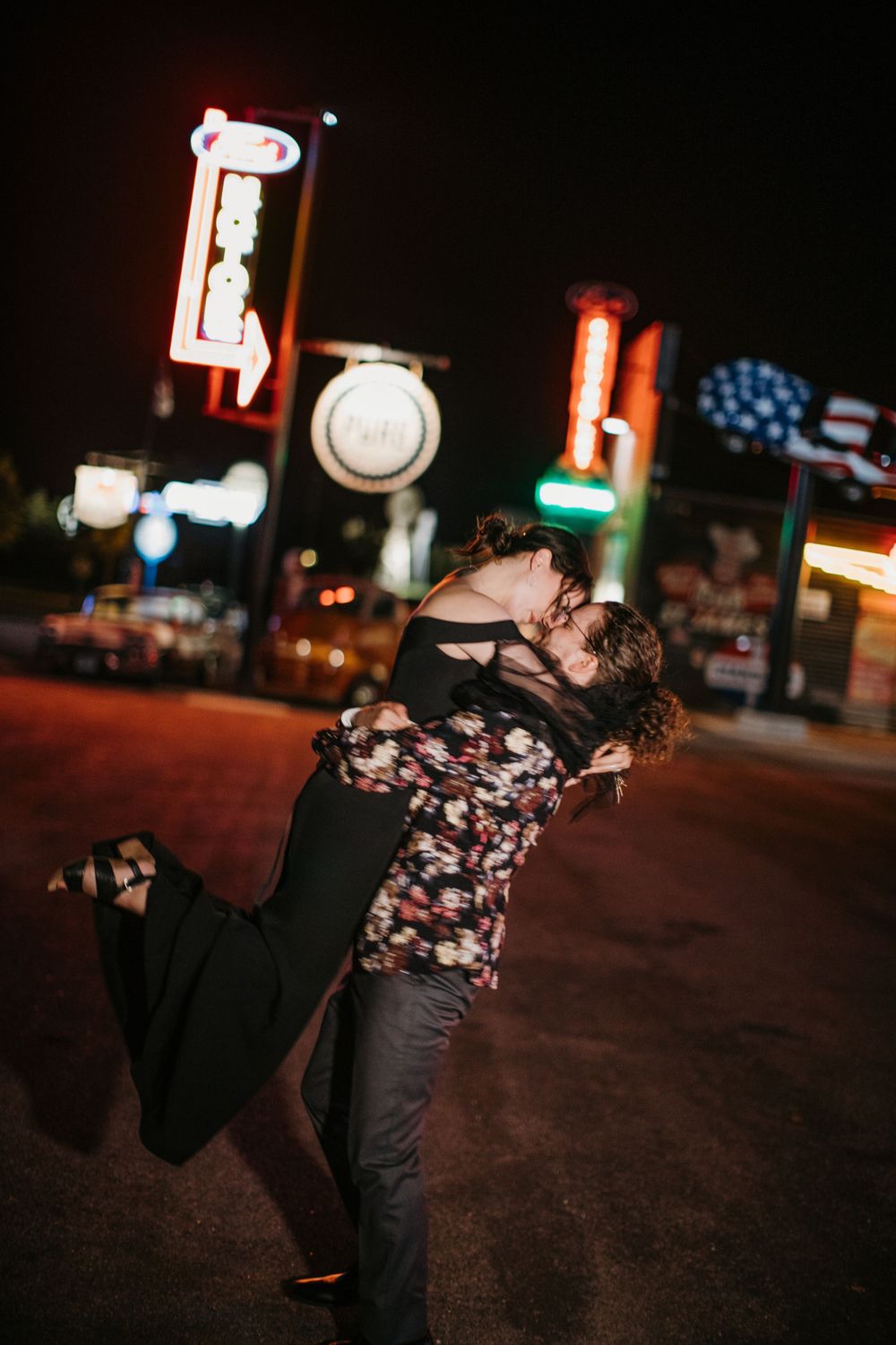 Couple sharing a romantic kiss as groom lifts bride near colorful neon signs at night.