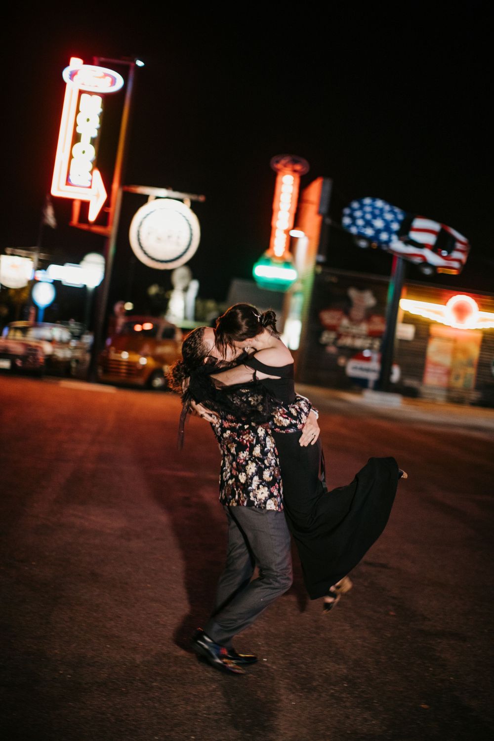 Couple embracing and kissing on a street surrounded by vibrant neon signs at night.