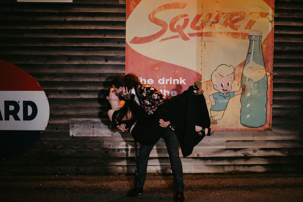 Couple sharing a romantic dip kiss in front of a vintage Squirt soda advertisement sign at night.