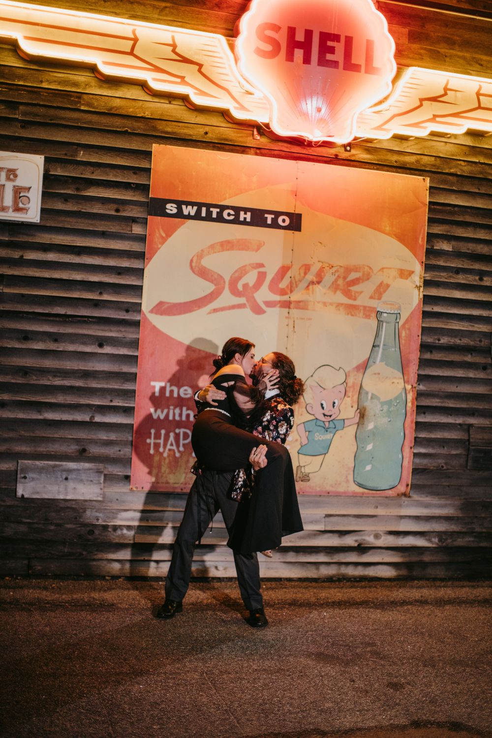Couple embracing and kissing in front of a vintage Squirt soda sign under a Shell neon light.