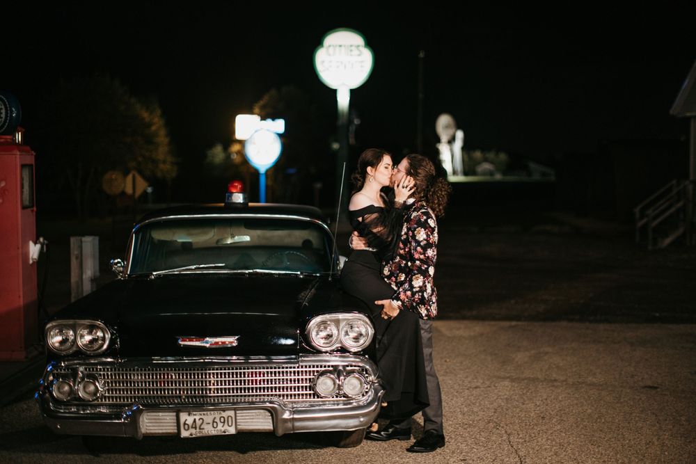 Couple kissing beside a classic black vintage car at a retro gas station at night.