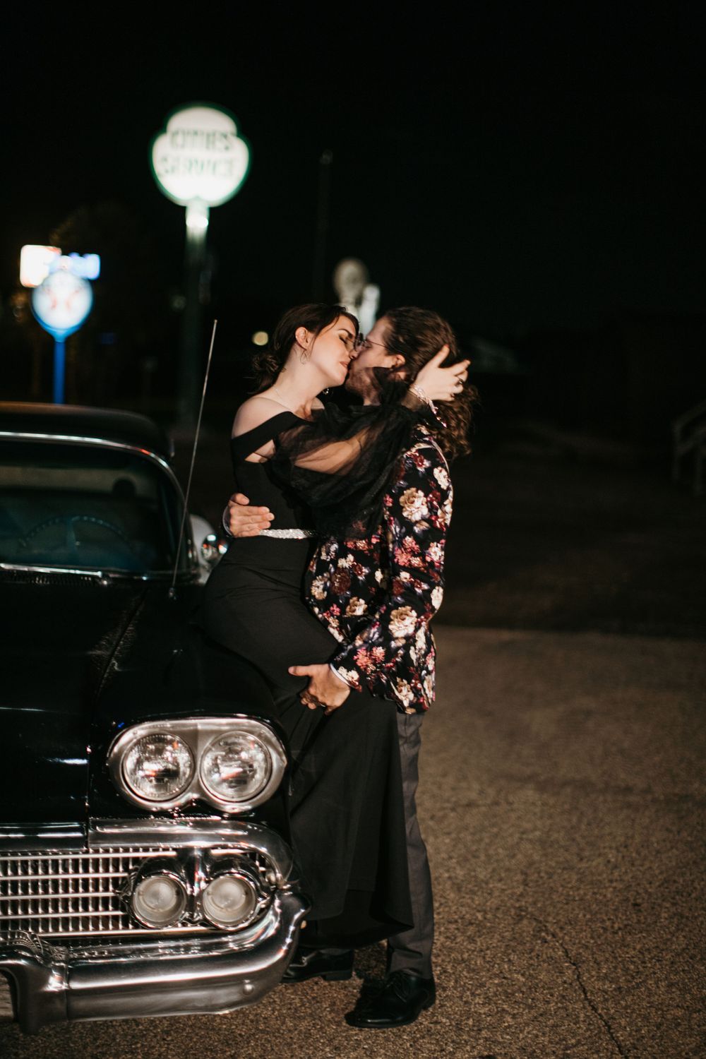Romantic couple kissing on the hood of a classic black vintage car at night.