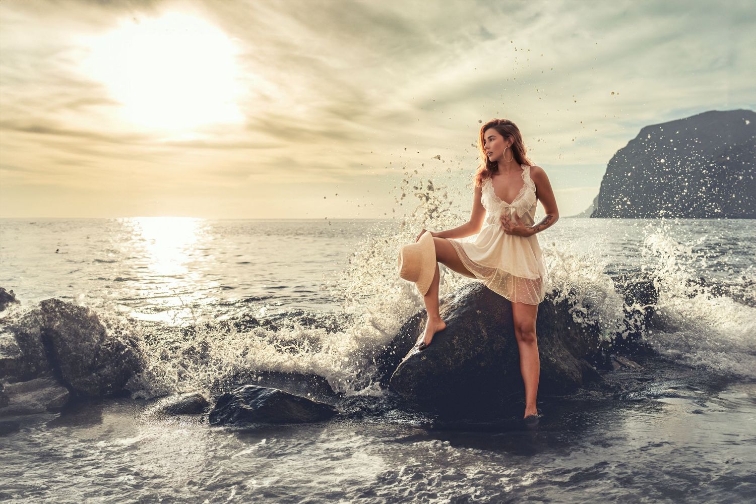 A white dress catches ocean spray as waves crash against coastal rocks at sunset.