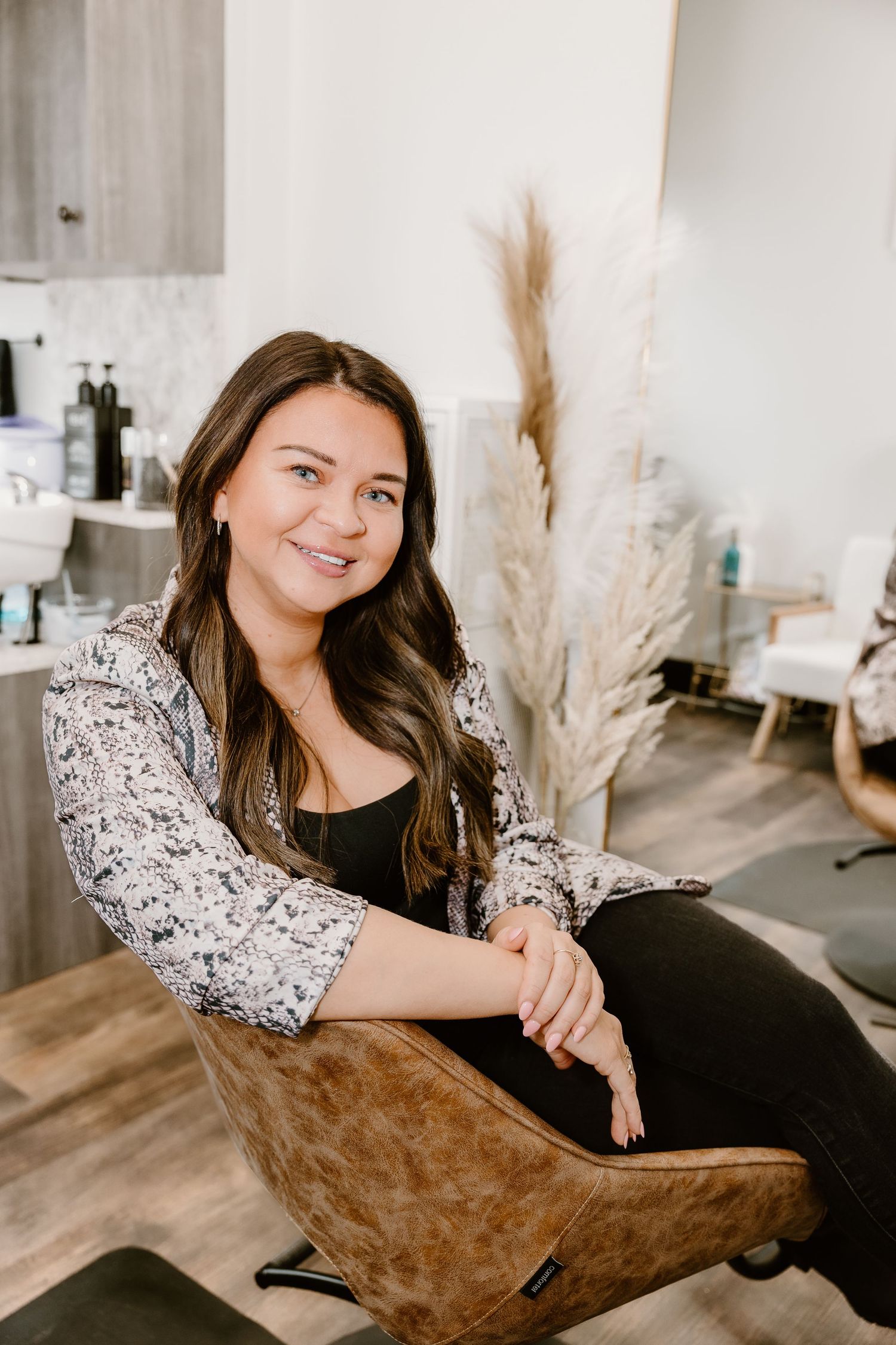 A hairstylist in snakeskin print blazer relaxes in a brown salon chair with pampas grass decor.