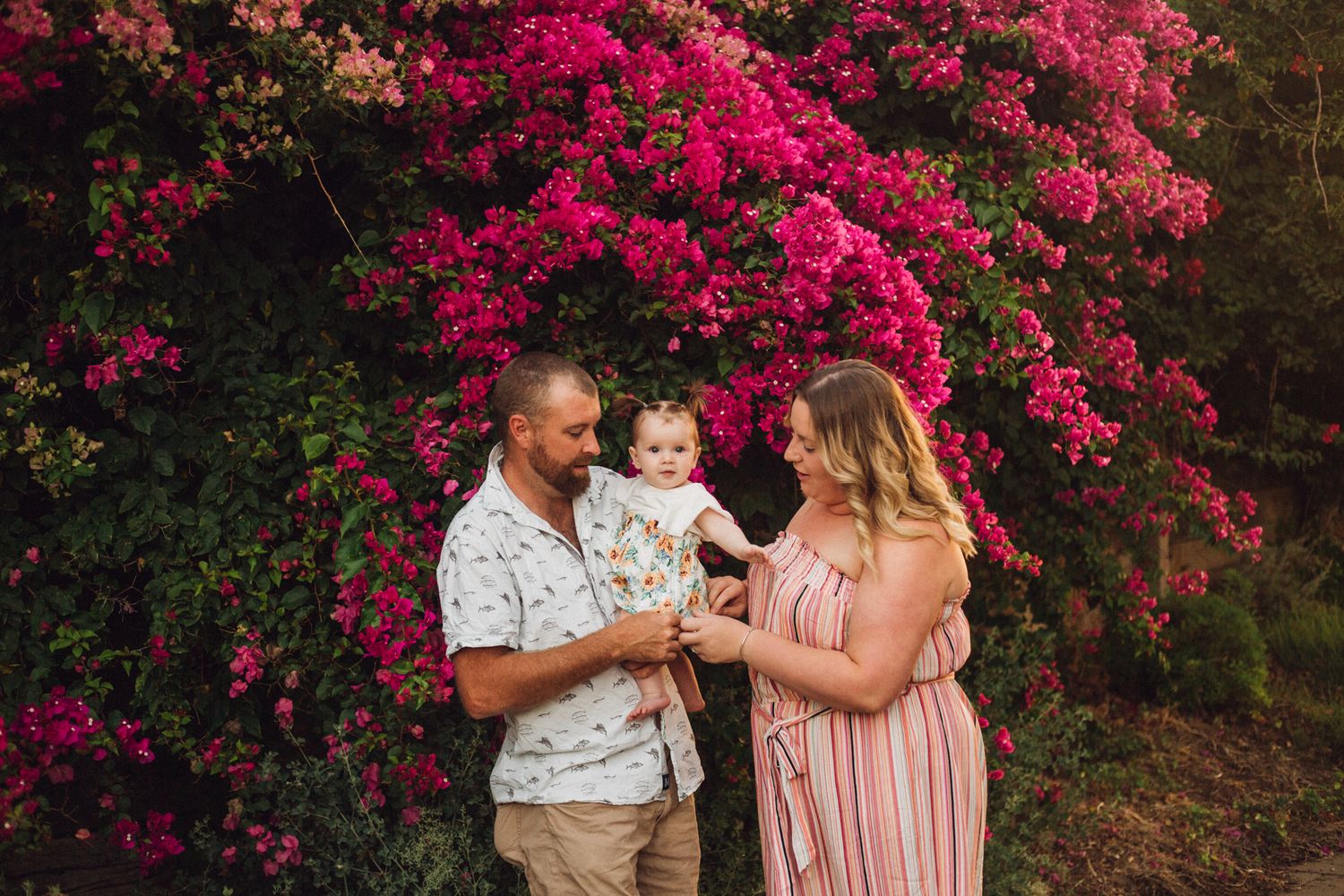 Dani and her husband holding their daughter infront of a large amount of pink flowers