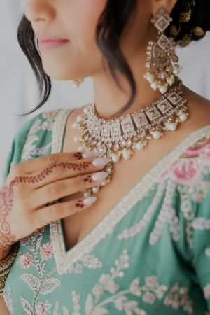 Close-up of diamond and pearl necklace with mehndi adorned hands on mint green embroidered fabric.