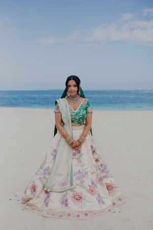 Bride wearing floral lehenga stands on white sandy beach against turquoise ocean backdrop.