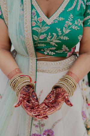 Close-up of henna adorned hands with gold bangles and embroidered green and white outfit.