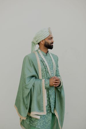 Man in mint green sherwani and turban with pearl necklaces against light background.
