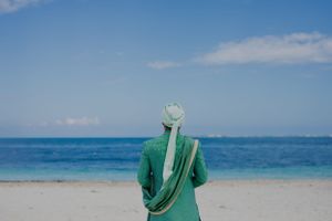 A person in green traditional attire stands on a beach overlooking turquoise ocean waters.