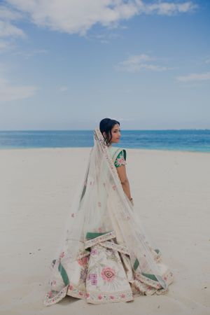 Bride in white floral lehenga poses gracefully on beach with flowing veil.