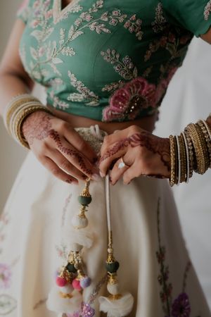Close-up of hands with mehndi holding decorative tassels against floral embroidered fabric.