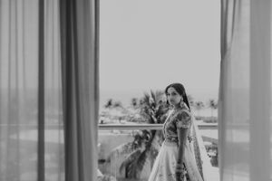 Black and white portrait of bride in traditional attire on balcony with palm trees in background.