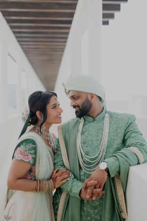A couple in mint green and ivory wedding attire sharing a tender moment in a white hallway.