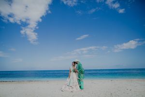 A couple in white and green wedding attire embracing on a pristine beach.