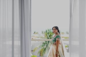 Bride in mint green traditional dress standing by curtained window with tropical view.
