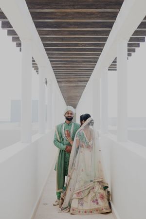 A couple in elegant green and cream traditional outfits posing in a modern white hallway.