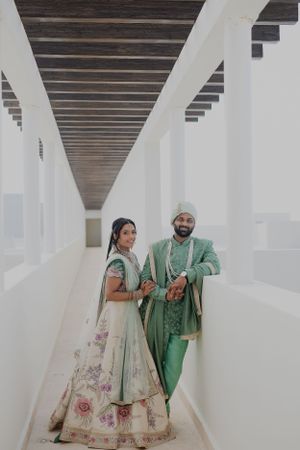 A couple in traditional Indian wedding attire poses in a modern white corridor.